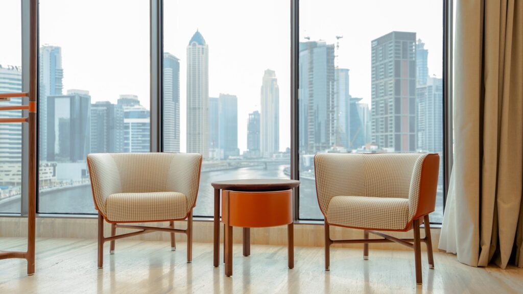 Modern lounge chairs beside floor-to-ceiling windows overlooking a Dubai Marina-style waterfront skyline, representing Al Fattan Marine Towers' JBR-Marina border setting