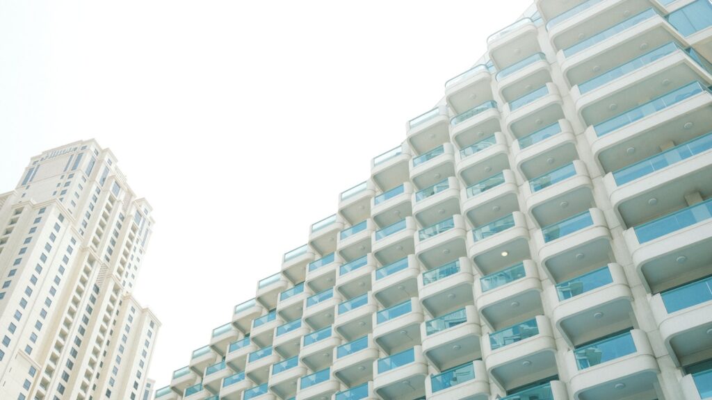 Low-angle view of a curved white residential tower with rows of glass balconies against a hazy sky, with a second cream tower visible in the background, representing modern high-rise architecture typical of DAMAC Hills