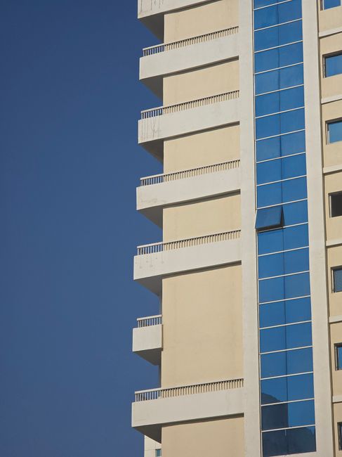 Close-up angled view of a contemporary high-rise facade with cream concrete balcony bands and a blue reflective glass curtain-wall section, representing Binghatti Corner in JVC, Dubai