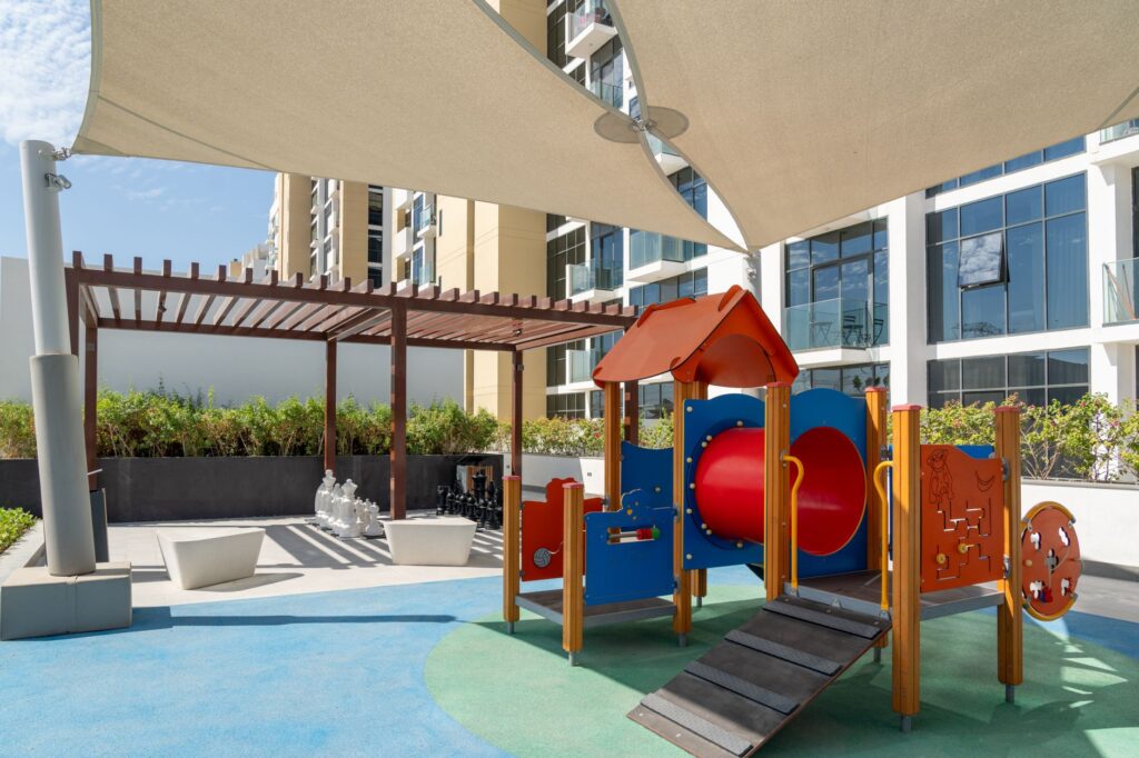 Shaded children's play structure with sail-shade canopy and landscaped greenery, with clean completed mid-rise residential buildings behind, representing Binghatti Onyx in JVC, Dubai