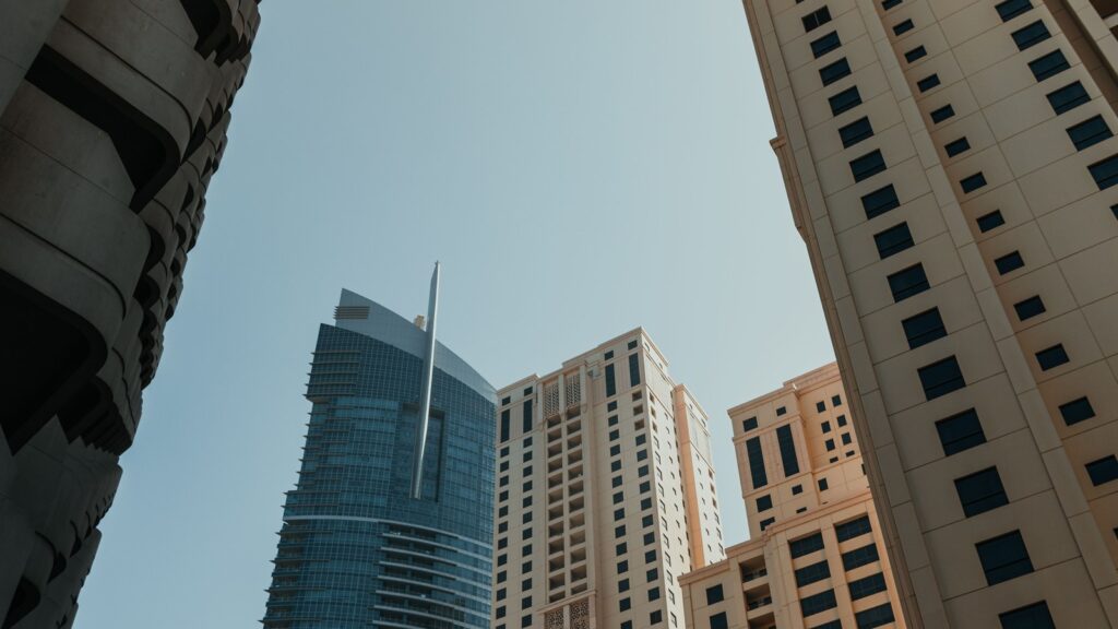 Low-angle view of modern Dubai high-rise apartment towers with a glass tower spire against a clear blue sky, representing the high-rise residential architecture typical of DAMAC Hills' Carson complex