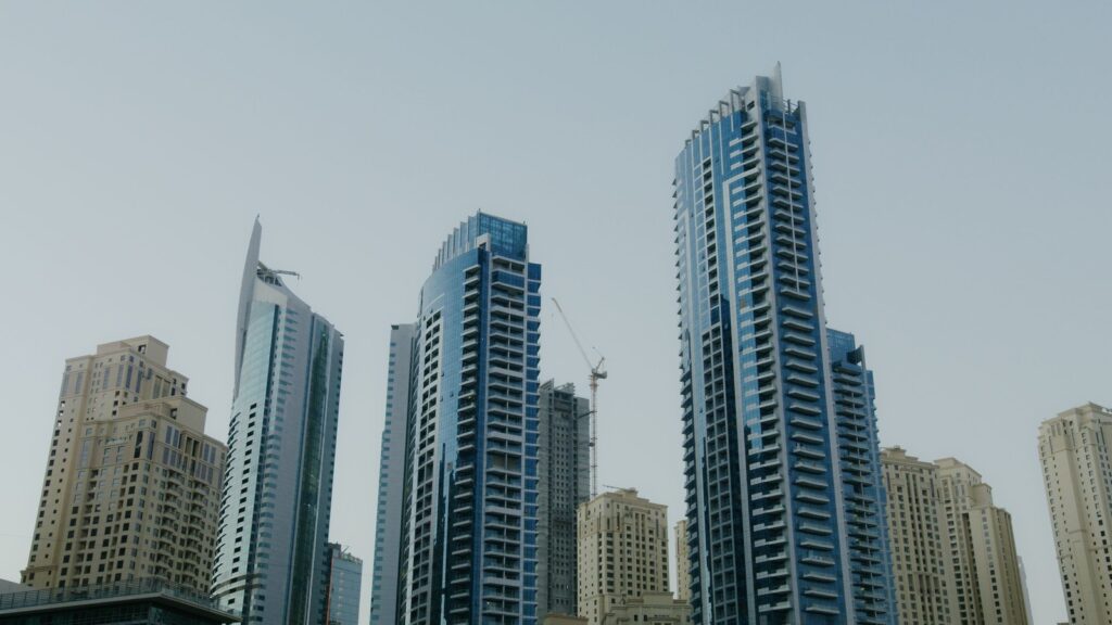 Low-angle view of tall blue-glass Dubai skyscrapers under a hazy sky with a distant construction crane, representing the fast-developing high-rise architecture typical of Dubai Creek Harbour