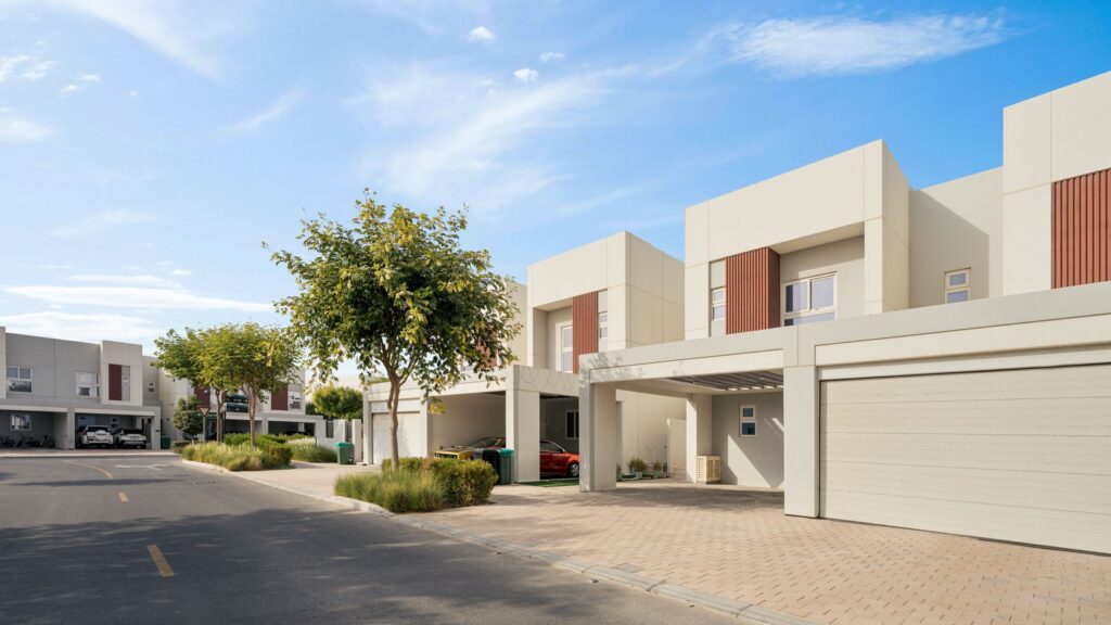 Row of modern cream-colored two-storey townhouses with covered parking and a street-side tree under a clear blue sky, representing the low-rise gated-community architecture typical of DAMAC Hills 2