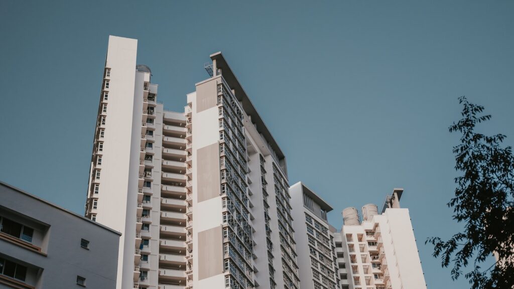 Low-angle view of a modern white residential apartment tower with rows of balconies against a clear blue sky and a small tree in the foreground, representing the residential architecture typical of Jumeirah Village Circle