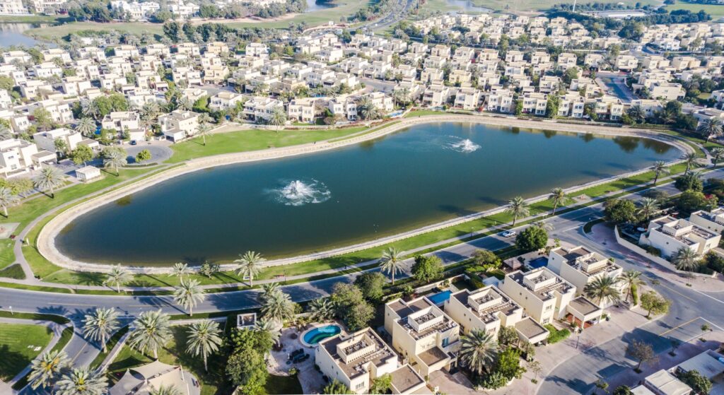 Aerial view of a landscaped lake with fountains surrounded by villa rooftops in a Dubai suburb