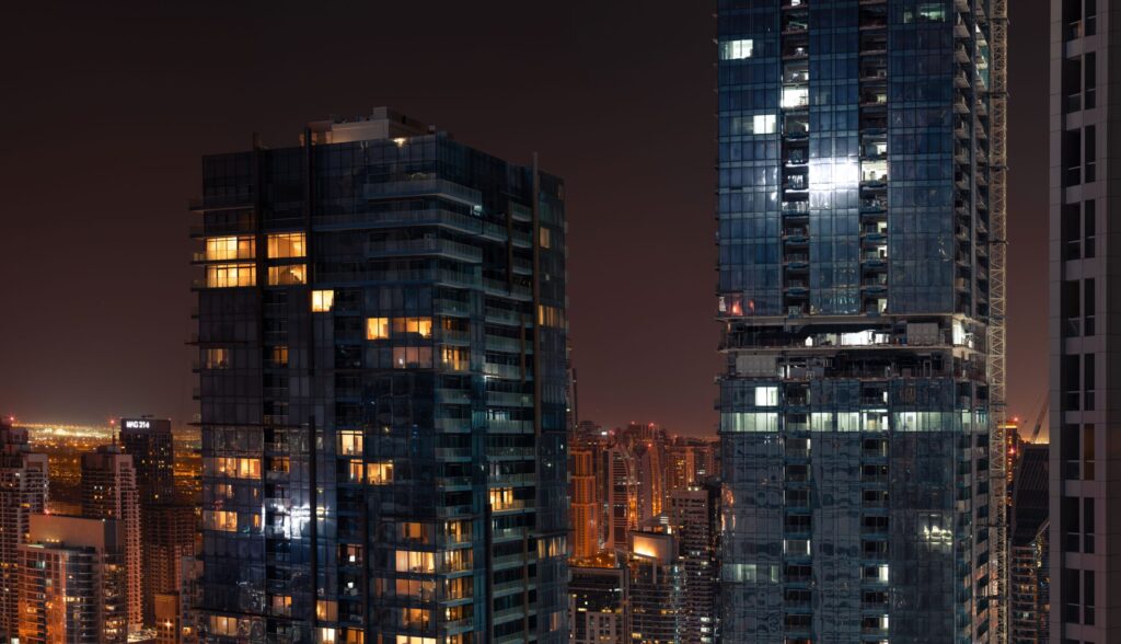 Two glass residential towers with warmly lit windows at night, a construction crane visible