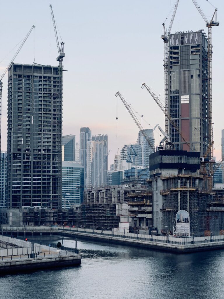 Two Dubai high-rise towers under construction with cranes above a waterfront skyline