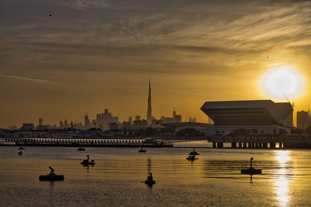 Burj Khalifa silhouette at golden-hour sunset across open water with the Museum of the Future's elliptical form visible