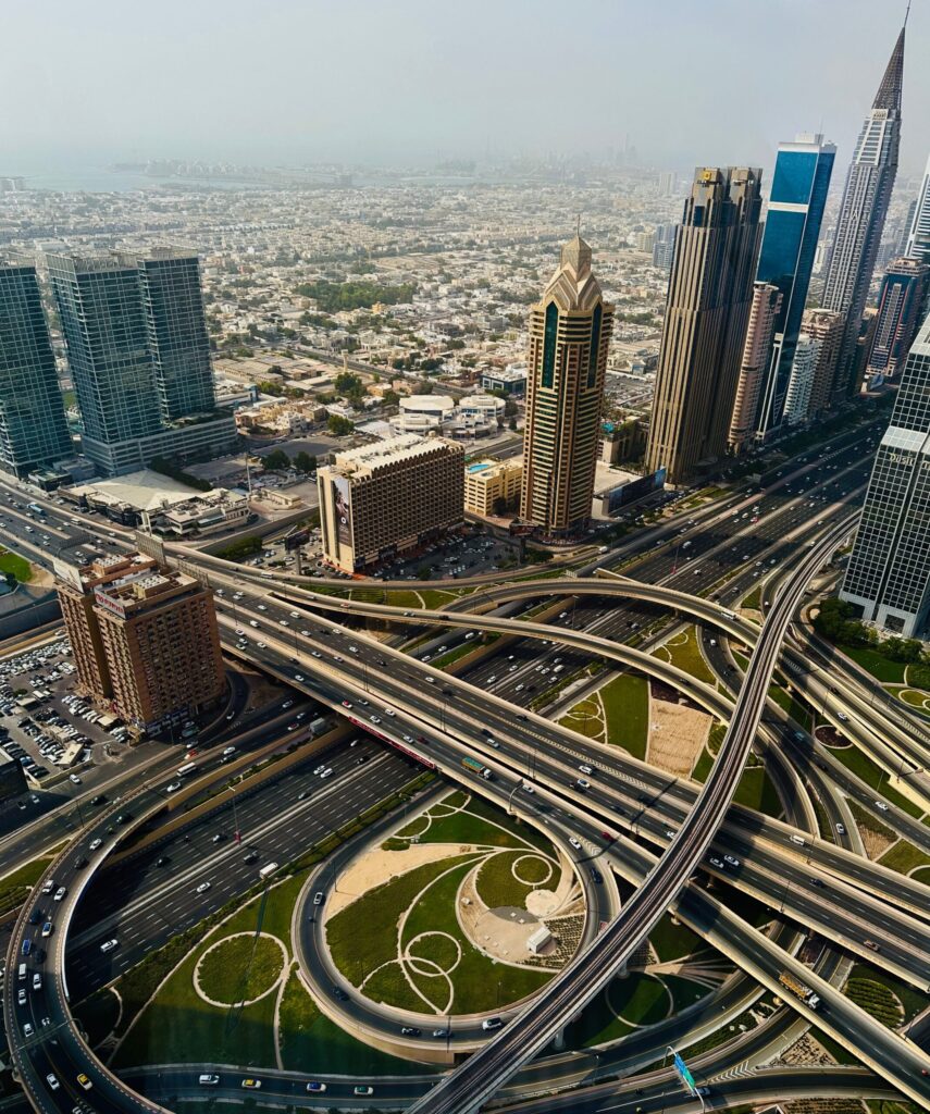High-angle aerial view of a Sheikh Zayed Road interchange in Dubai with a distant tower cluster in soft haze