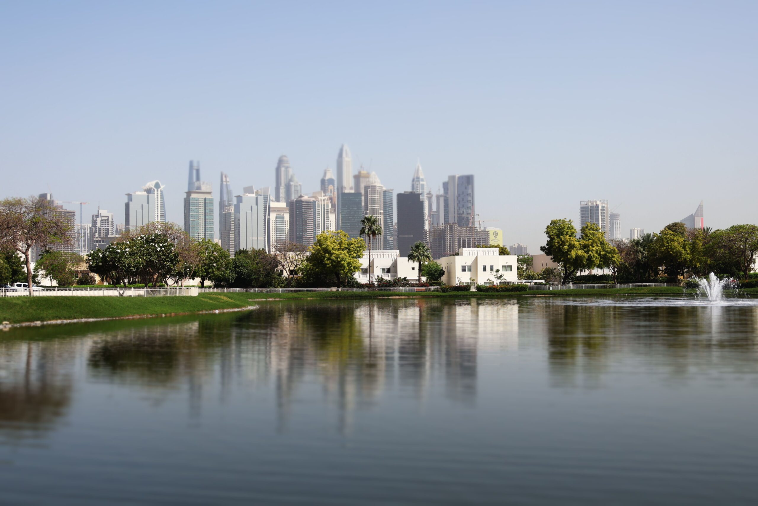 Landscaped community lake with a working fountain and low-rise white and cream villa rooftops framed by palm trees, with the Dubai Marina and JLT tower skyline reflected in the water, representative of The Springs' Emirates Living community, Dubai