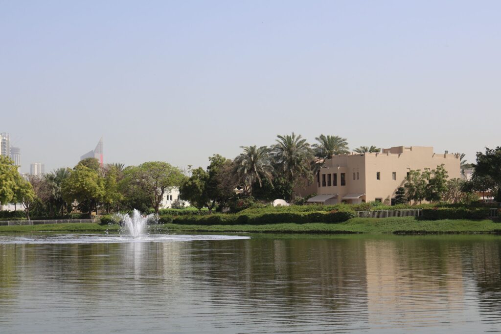 Landscaped community lake with fountain and a traditional beige-stucco villa with palm trees, Dubai Marina and JLT tower skyline visible in the distance, representative of The Springs' Emirates Living community, Dubai