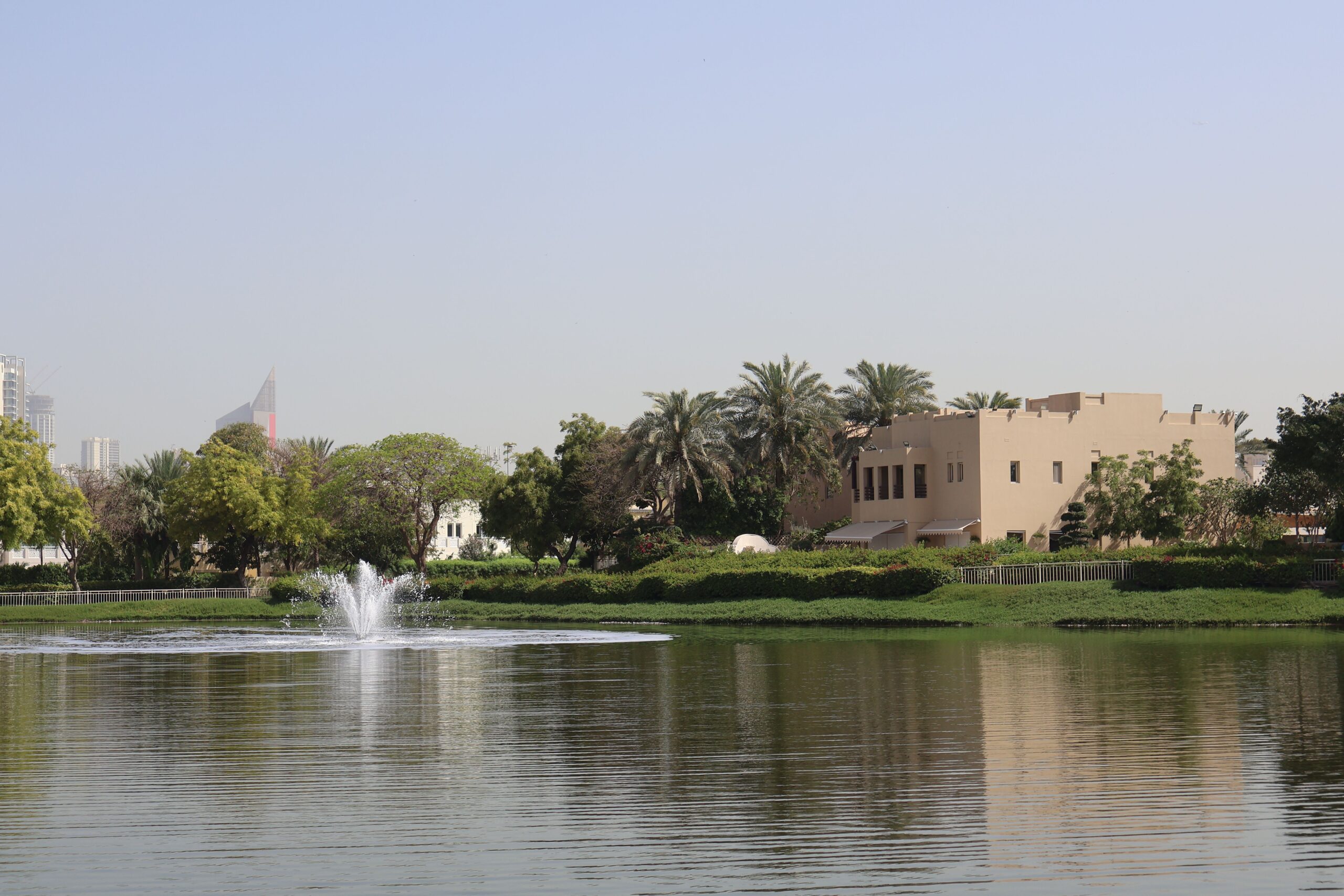 Landscaped community lake with fountain and a traditional beige-stucco villa with palm trees, Dubai Marina and JLT tower skyline visible in the distance, representative of The Springs' Emirates Living community, Dubai