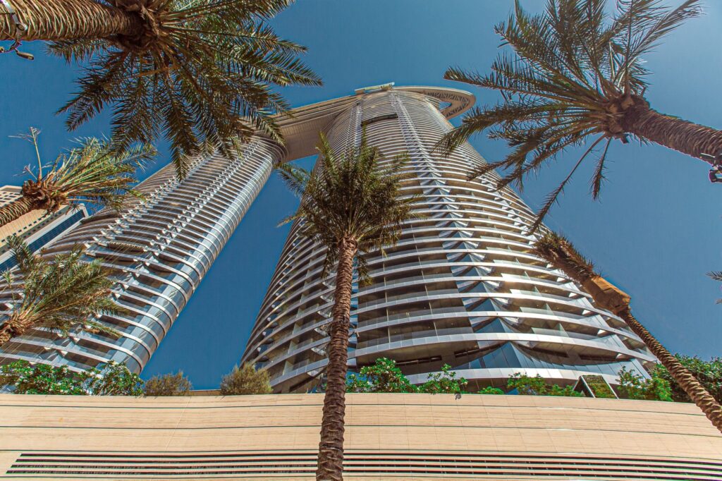Low-angle view of two curved, ribbed-glass high-rise towers framed by palm fronds against a clear blue sky, representative of The Walk, JBR, Dubai