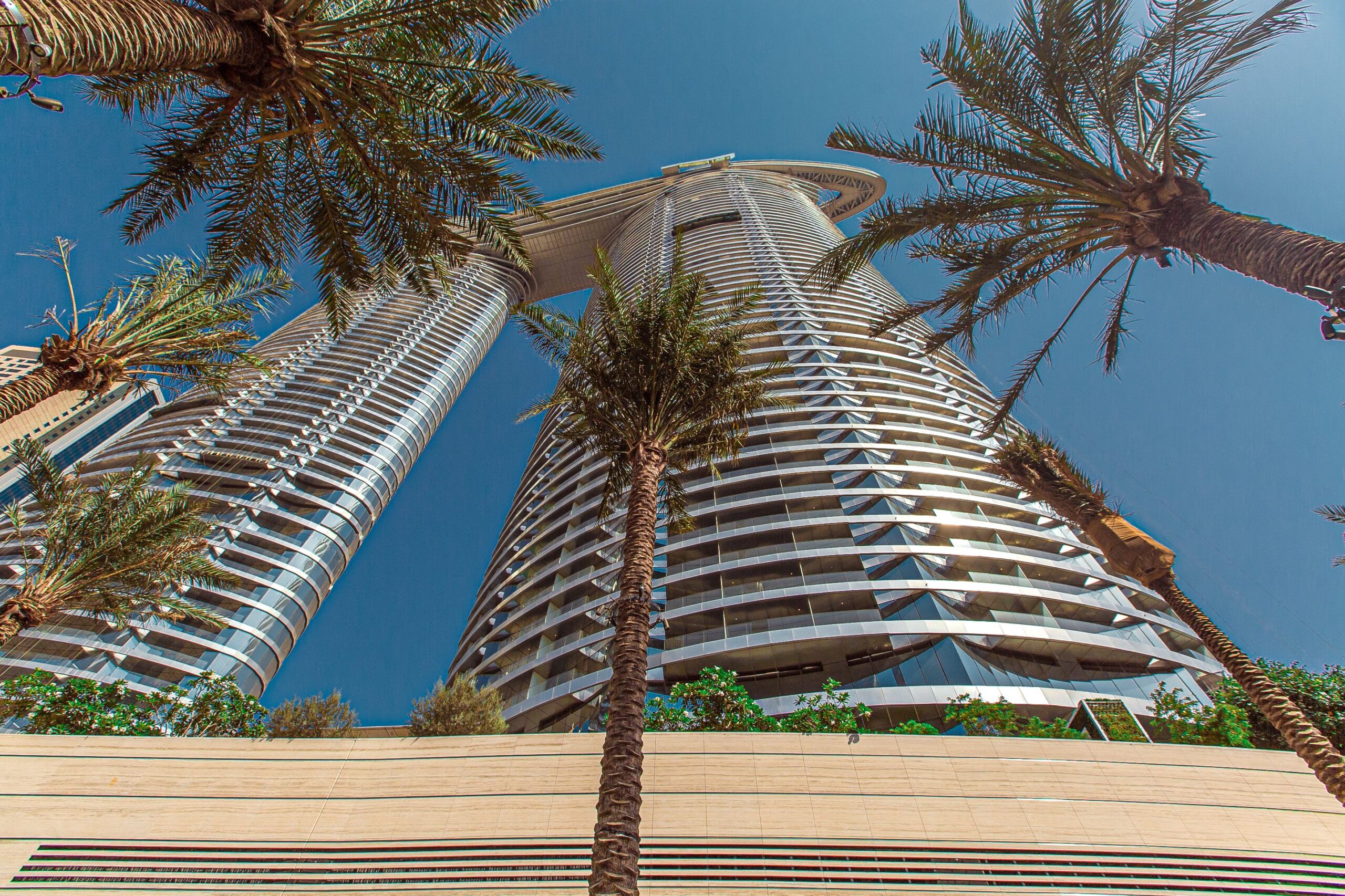Low-angle view of two curved, ribbed-glass high-rise towers framed by palm fronds against a clear blue sky, representative of The Walk, JBR, Dubai
