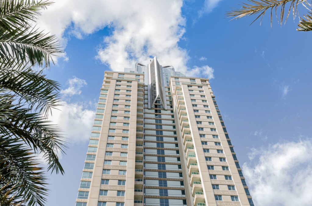 Low-angle view of a modern cream high-rise tower with vertical facade banding and glass balconies against a blue sky, framed by palm fronds