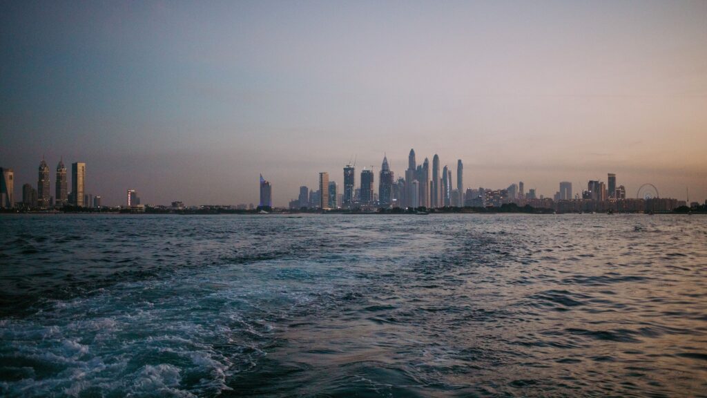 Dubai Marina and JBR skyline viewed across calm sea water at dusk, with towers silhouetted against a soft sunset sky, representing Shams 1's coastal JBR setting