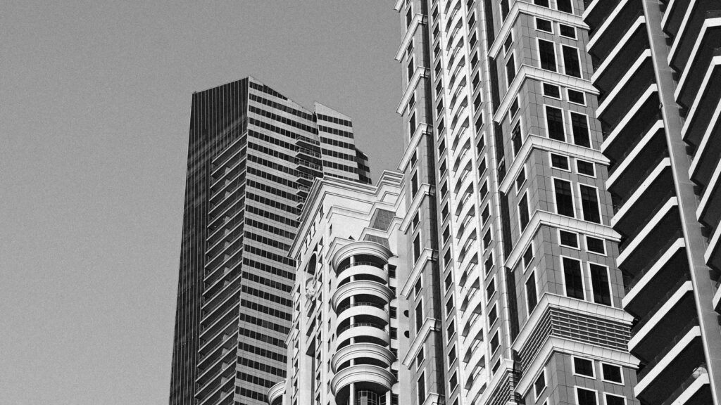 Black and white low-angle view of layered Dubai high-rise towers with glass and concrete facades against a clear sky, representing Shams 2's JBR tower setting