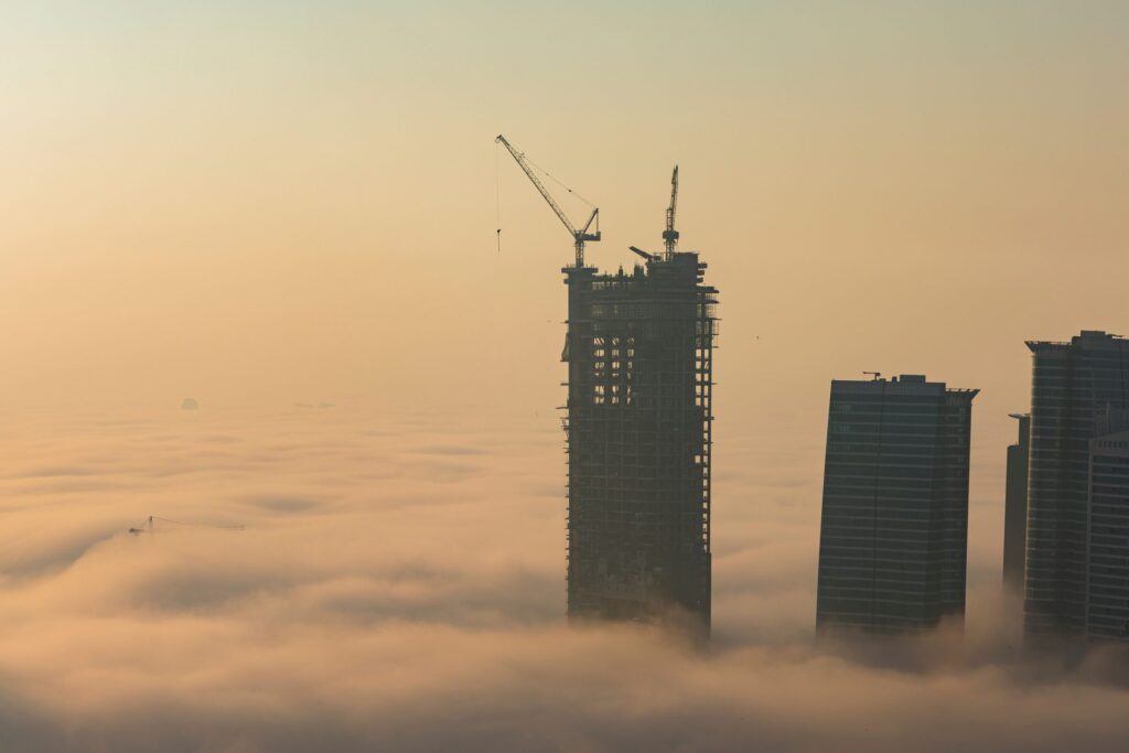 Aerial dawn view of Dubai skyscrapers emerging from thick fog with a construction crane visible atop one tower, representative of Sobha Hartland II's early-stage construction