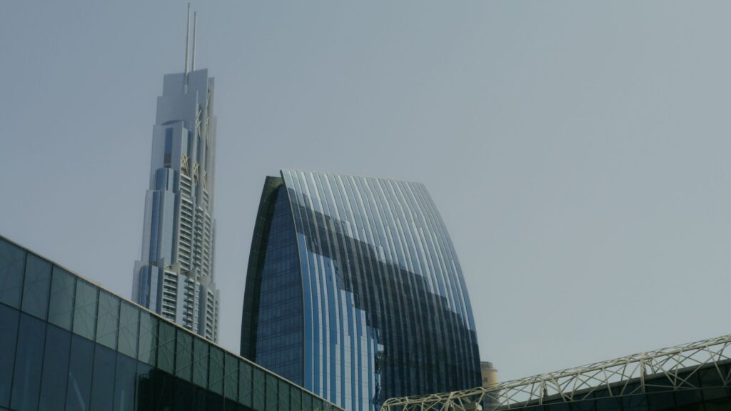 Low-angle view of distinctive Dubai glass skyscrapers including a curved-facade tower and a slender spire tower against a hazy sky, representing the waterfront high-rise architecture typical of Emaar Beachfront