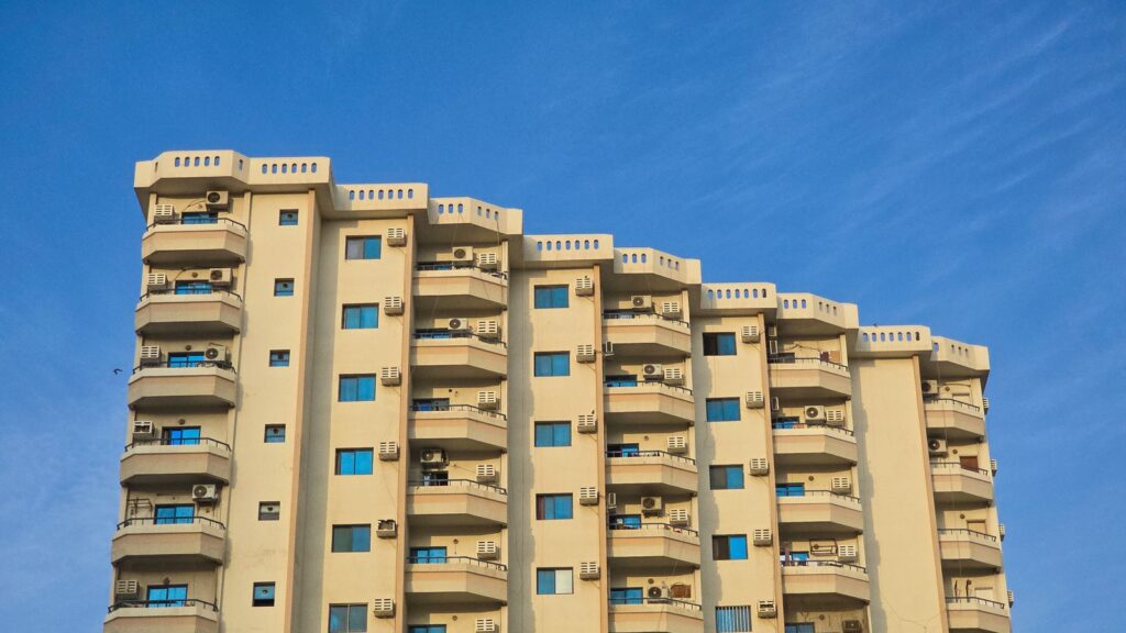 Low-angle view of a cream-colored modern residential apartment building facade with rows of balconies against a clear blue sky, representing the residential-tower architecture typical of Sobha Hartland