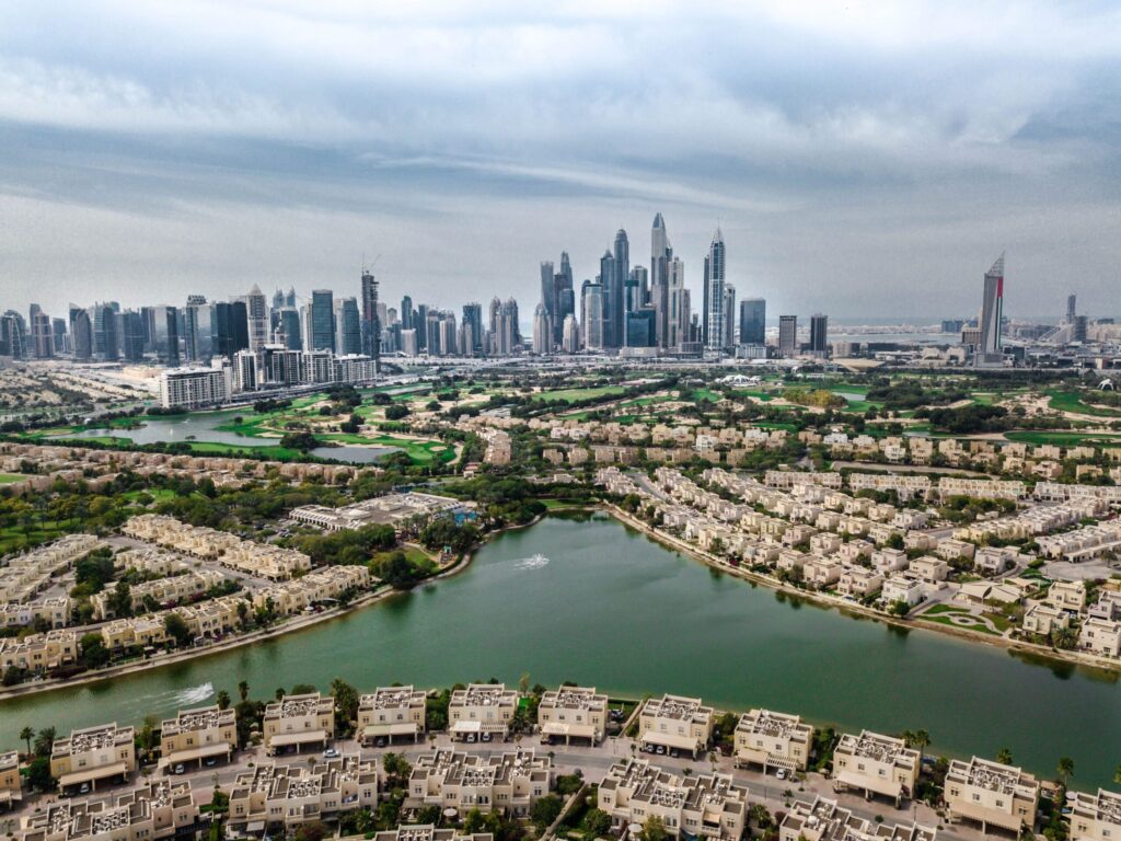 Aerial view of a Dubai villa community with lakes and a golf course, cream low-rise villa clusters wrapping around the water with the Dubai Marina skyline visible in the distance, representative of Springs 7, The Springs, Emirates Living