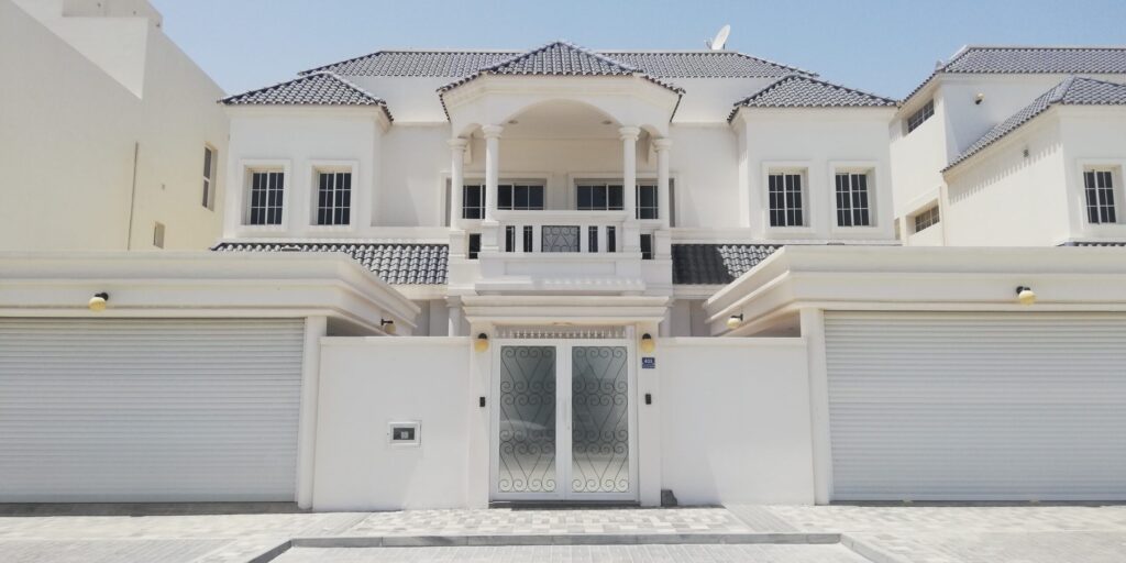 White Gulf-style two-storey villa with an arched entrance, dark tiled roof and roller-door garage near Indigo Ville 7, JVC