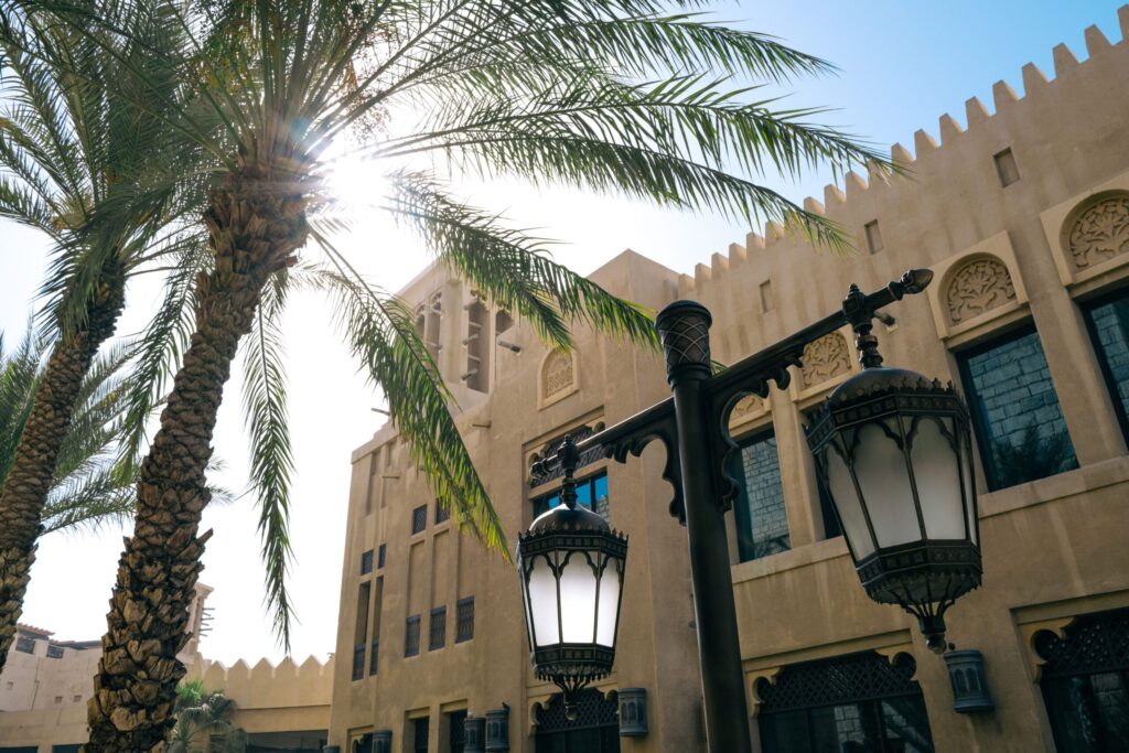 Sandstone Arabian-style building with ornate lanterns and wind-tower architecture behind a palm tree in Old Town, Downtown Dubai, near Reehan 5