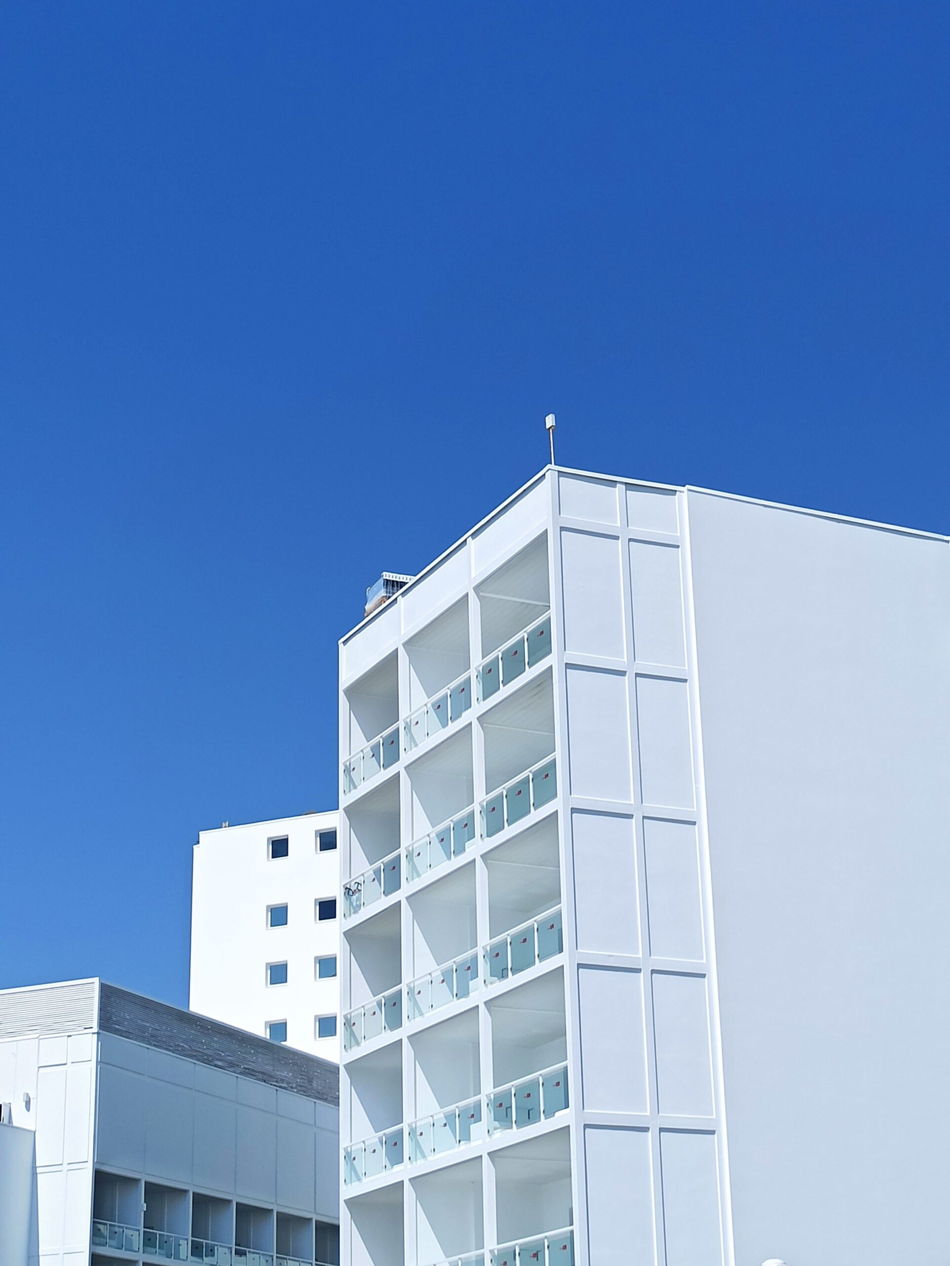 Clean white modern apartment building corner with glass balcony railings under a clear blue sky, representative of Binghatti Residences in Dubai Silicon Oasis, Dubai