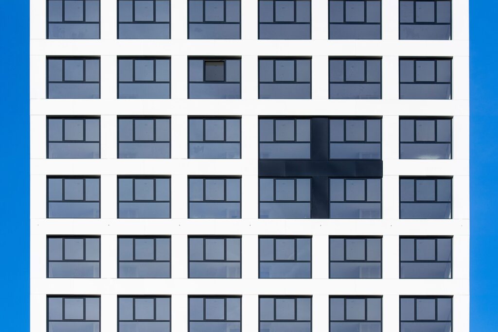 Close-up of a modern white apartment building facade with a uniform grid of dark-framed windows and a dark cross-shaped accent panel against a vivid clear blue sky, evocative of a Liwan, Dubai residential development