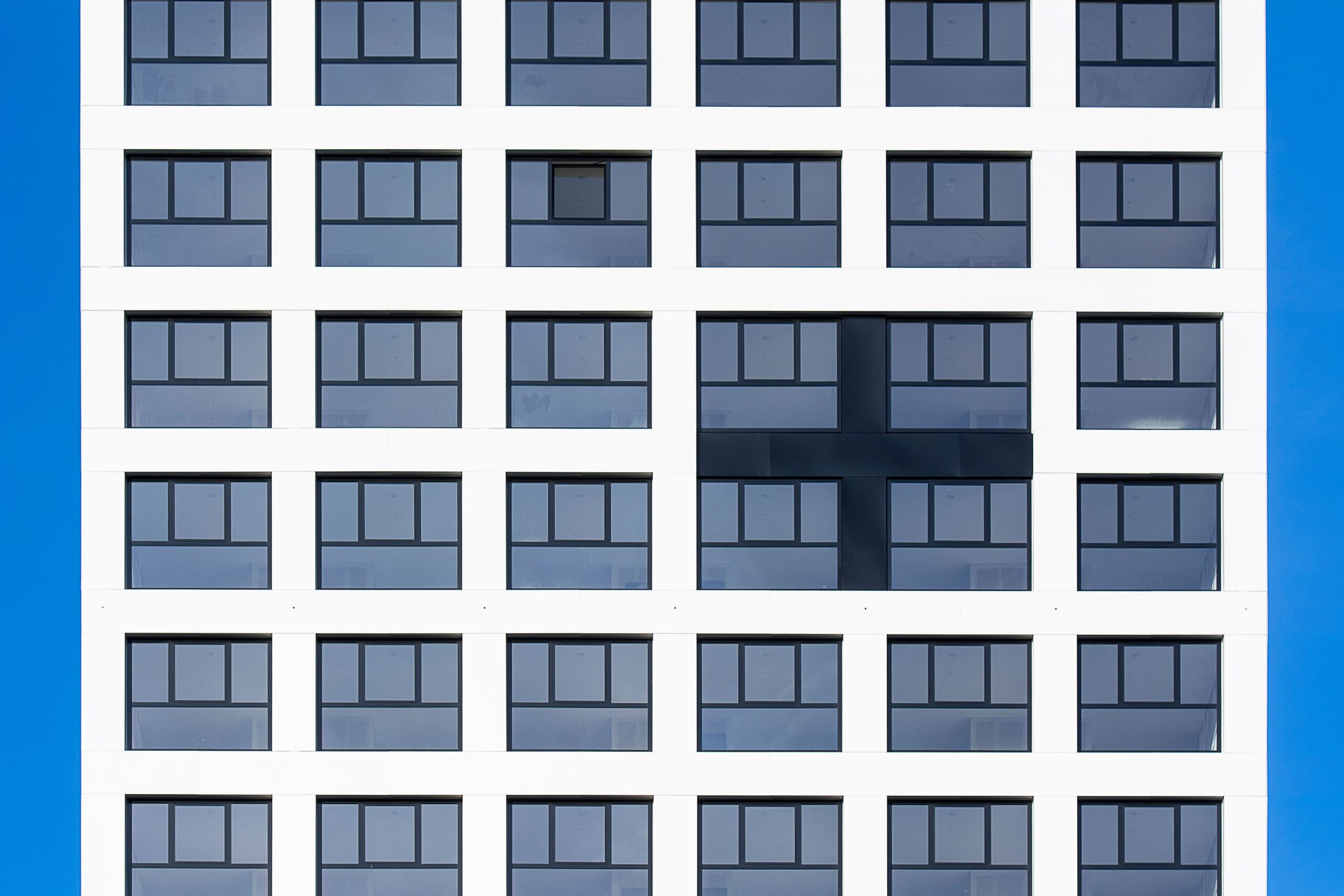 Close-up of a modern white apartment building facade with a uniform grid of dark-framed windows and a dark cross-shaped accent panel against a vivid clear blue sky, evocative of a Liwan, Dubai residential development