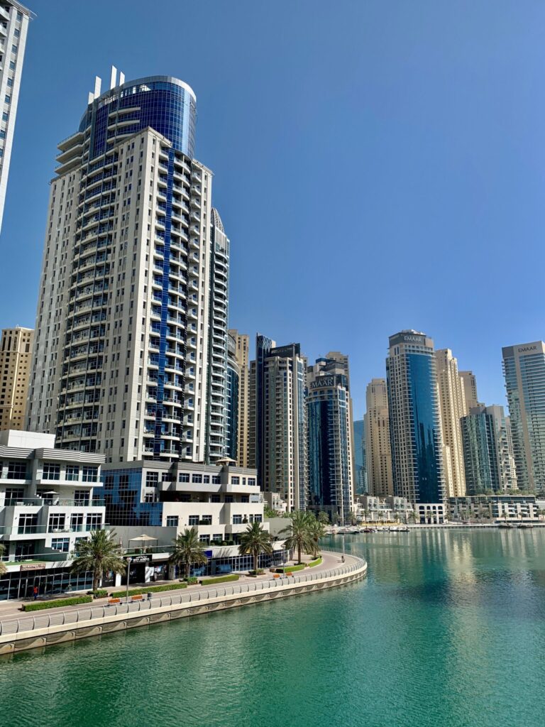 Daytime view of a curved waterfront residential tower beside a teal canal with palm trees, in a Dubai waterfront district similar to Sobha Hartland