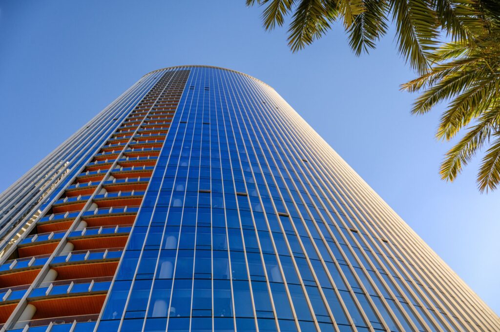 Low angle view of a curved blue glass high-rise tower against a clear sky with palm fronds in the foreground, Dubai