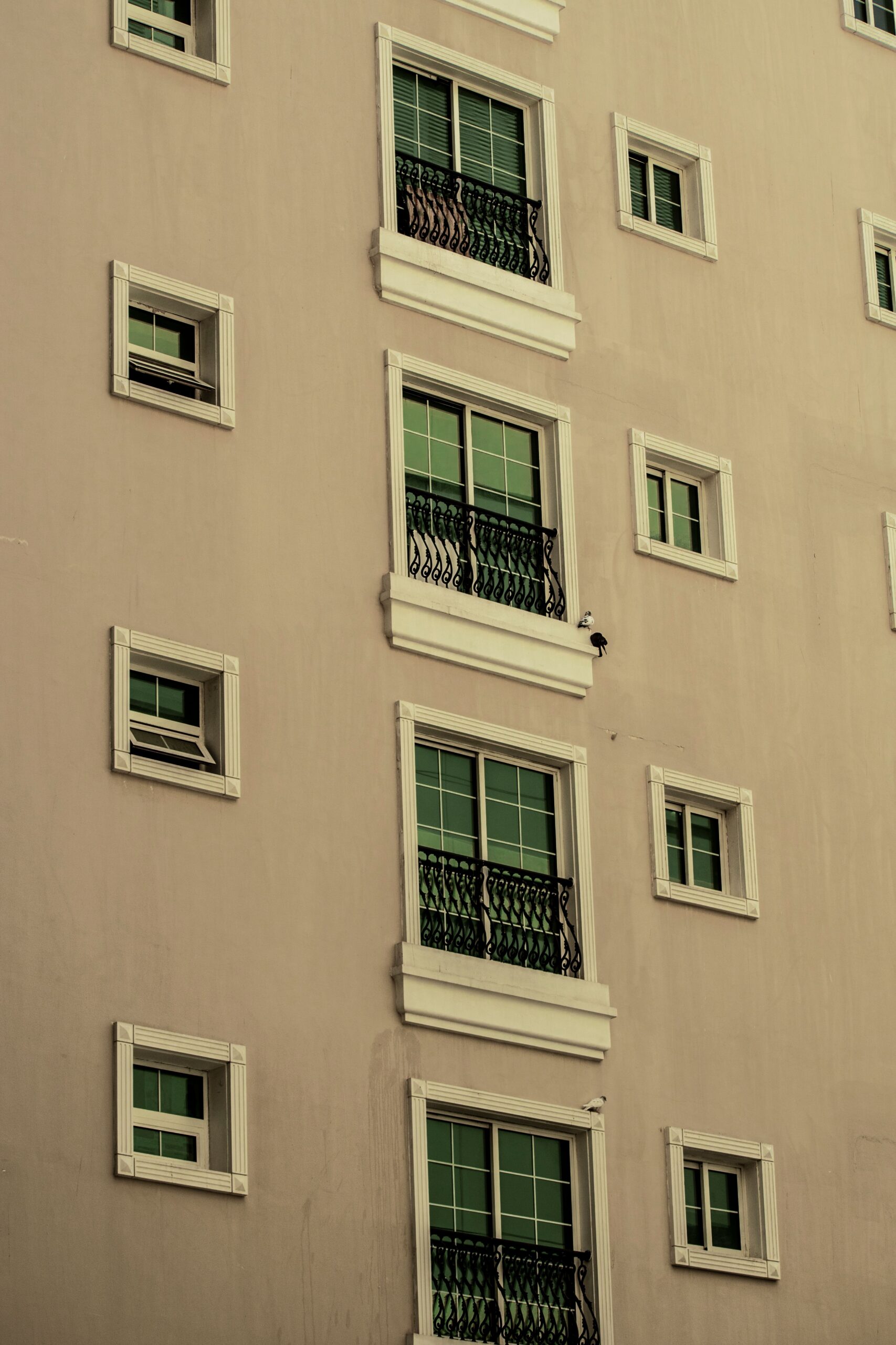 Symmetrical facade of a beige mid-rise Dubai apartment building with rows of windows and small wrought-iron Juliet balconies against a hazy sky, representative of The Crescent Tower A, Dubai Production City