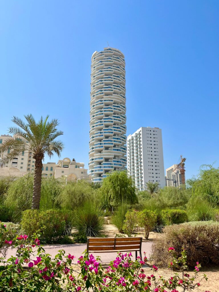 A distinctive round high-rise residential tower seen past palm trees and flowering bougainvillea under a clear blue sky, evocative of Socio's setting in Dubai Hills Estate