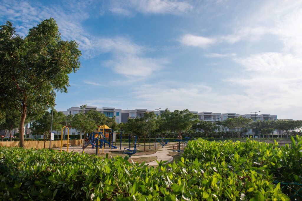 Children's play area with low-rise cream residential buildings and mature trees in the background under a clear blue sky, representative of the shared community amenities near Springs 4, The Springs, Emirates Living
