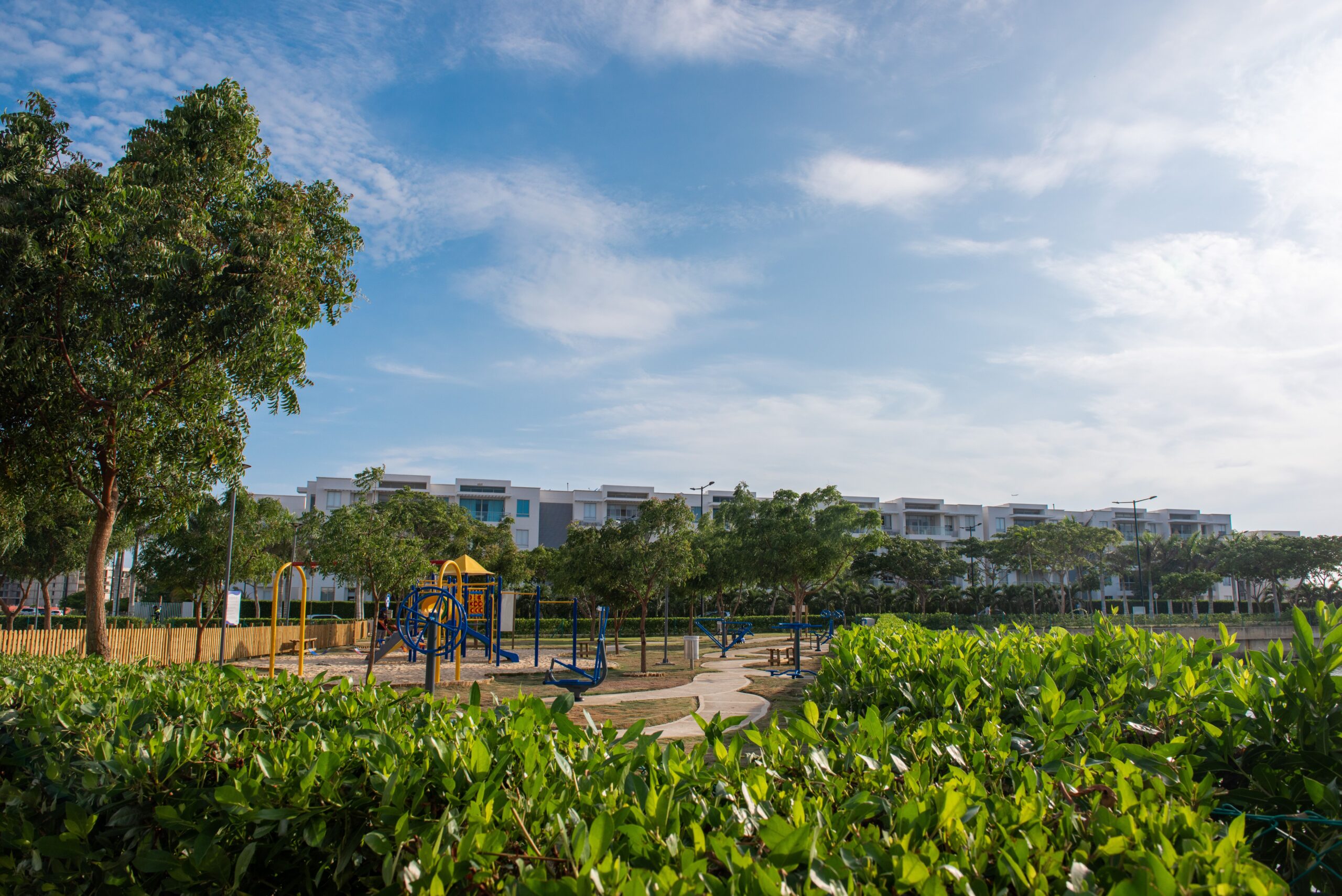 Children's play area with low-rise cream residential buildings and mature trees in the background under a clear blue sky, representative of the shared community amenities near Springs 4, The Springs, Emirates Living