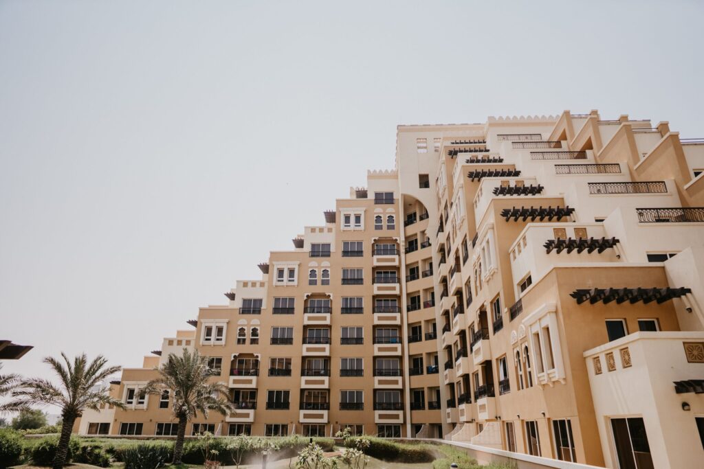 Cream and tan Arabic-style stepped mid-rise apartment building with arched windows and palm trees in the foreground under a clear blue sky in Dubai, representative of Azizi Star's residential architecture in Al Furjan