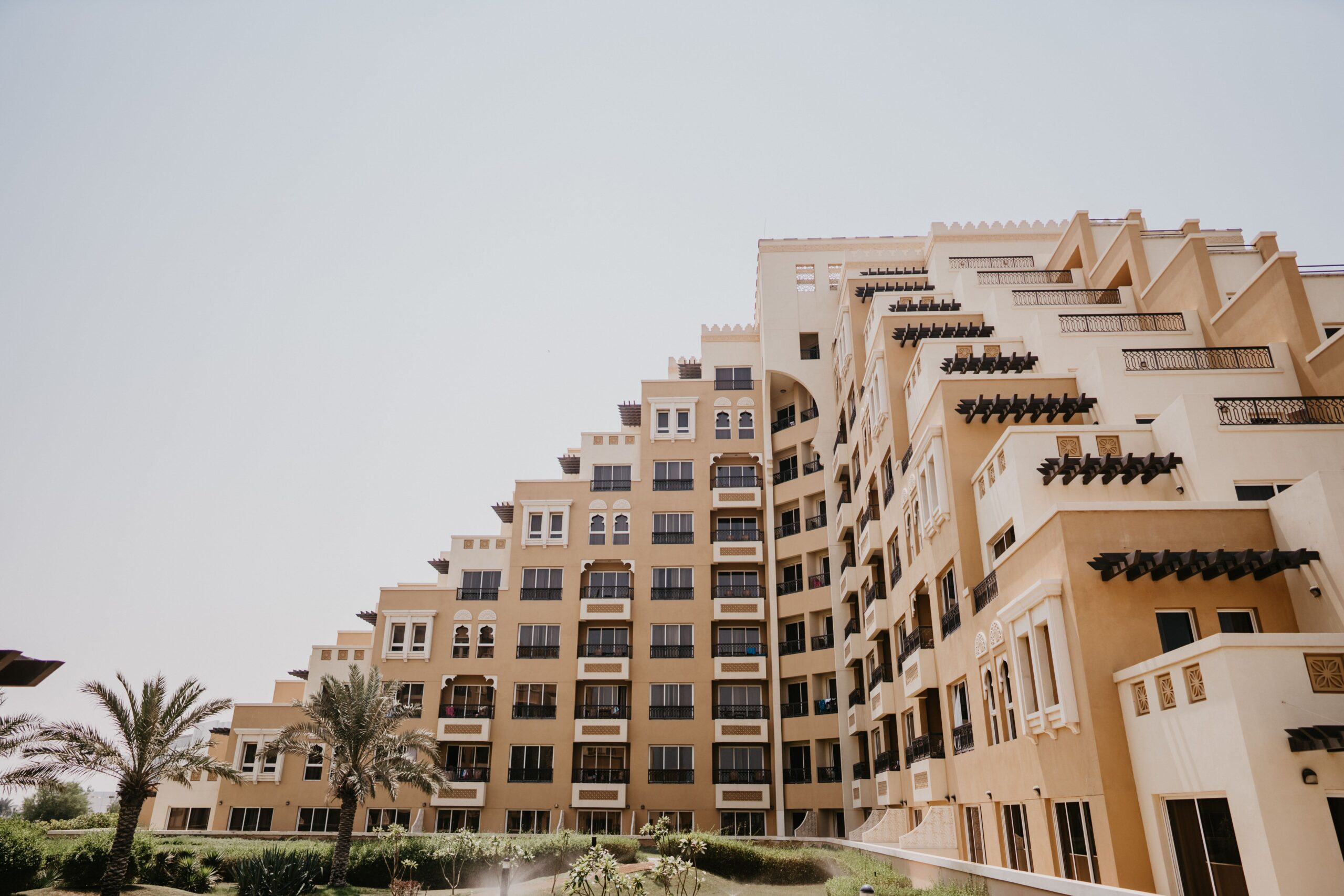 Cream and tan Arabic-style stepped mid-rise apartment building with arched windows and palm trees in the foreground under a clear blue sky in Dubai, representative of Azizi Star's residential architecture in Al Furjan