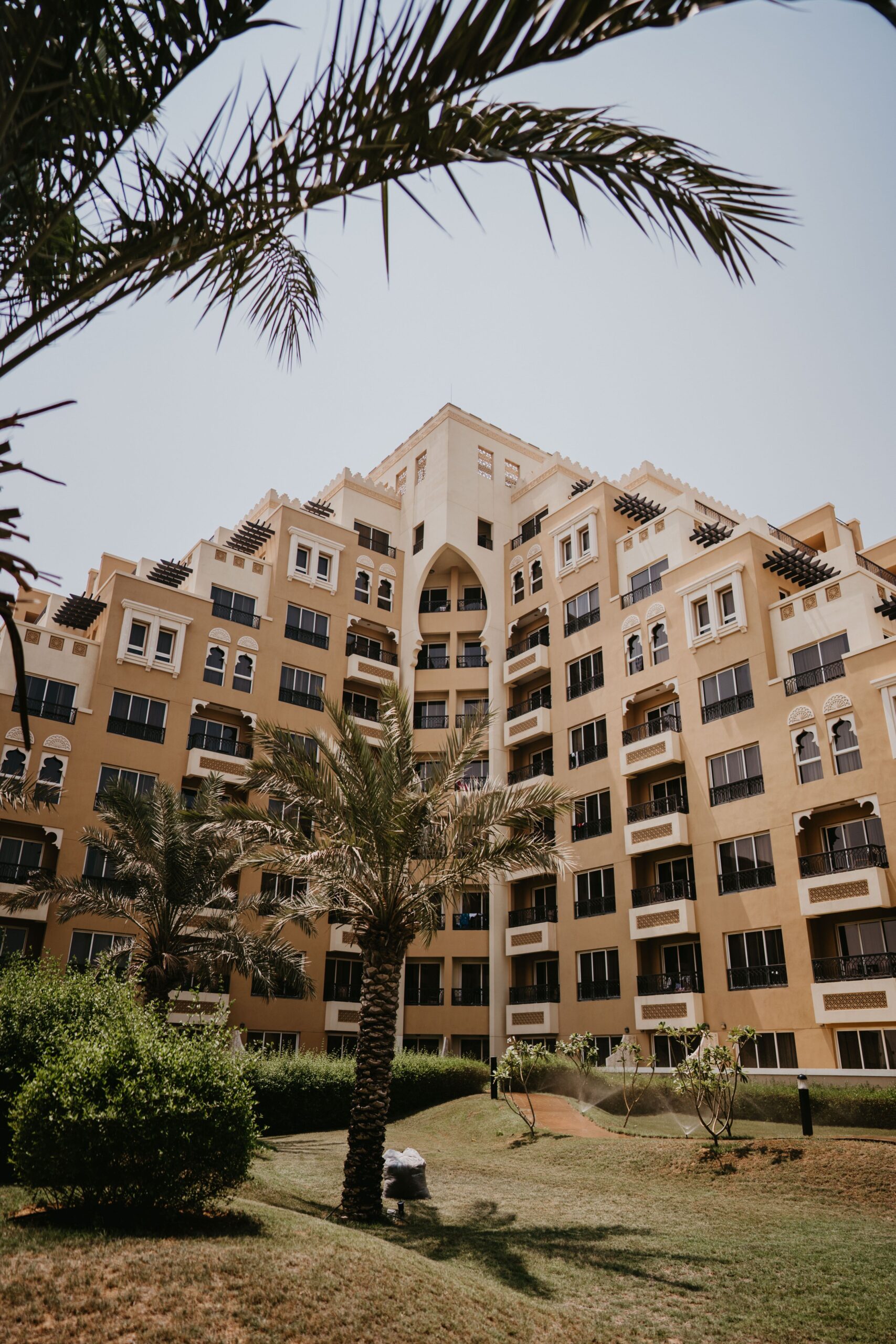 Low-angle view of a cream and tan Arabian-style mid-rise apartment building with arched mashrabiya window details and a palm tree in the foreground under a clear blue sky in Dubai, representative of Azizi Orchid's residential architecture in Al Furjan