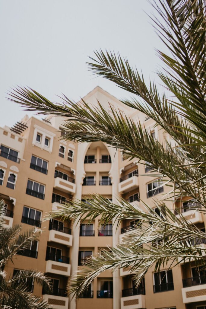 Upward view of a beige mid-rise Dubai apartment building with an arched Gulf-style balcony feature and palm fronds in the foreground, representative of Olivz by Danube's residential architecture in International City