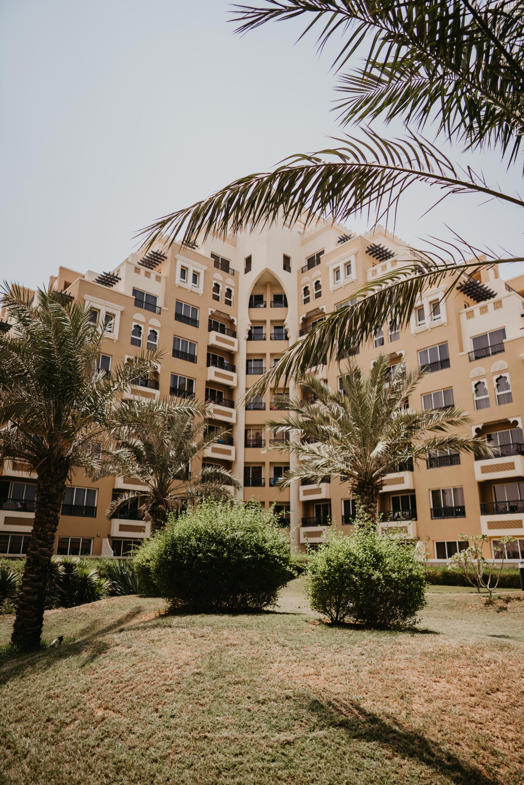 Cream and tan Arabic-style stepped mid-rise apartment building with arched windows and palm trees in the foreground under a clear blue sky in Dubai, representative of Azizi Plaza's residential architecture in Al Furjan