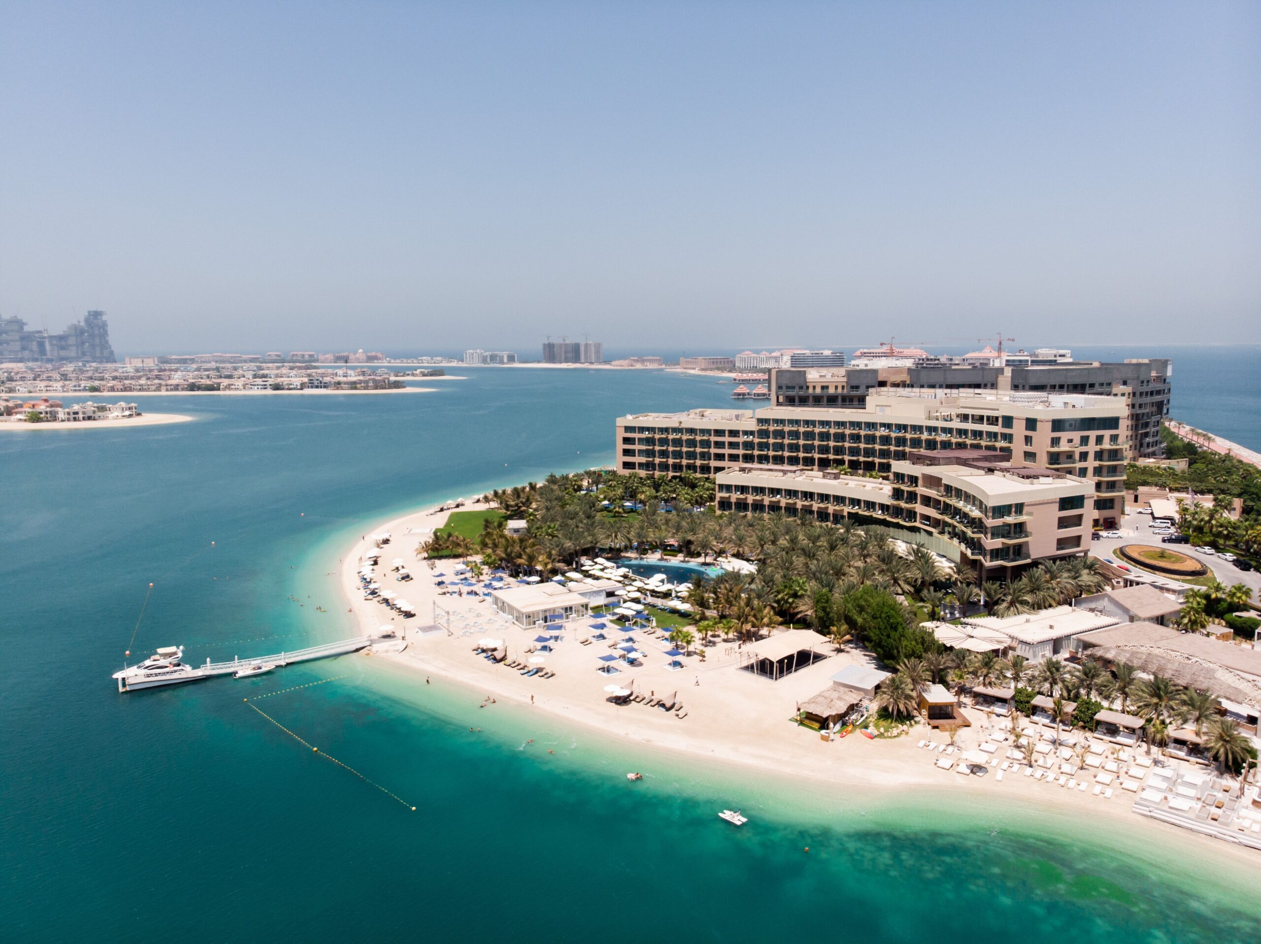 Aerial view of a mid-rise beachfront hotel and resort complex on a Palm Jumeirah beach curve, near Seabird Residence on Palm West Beach