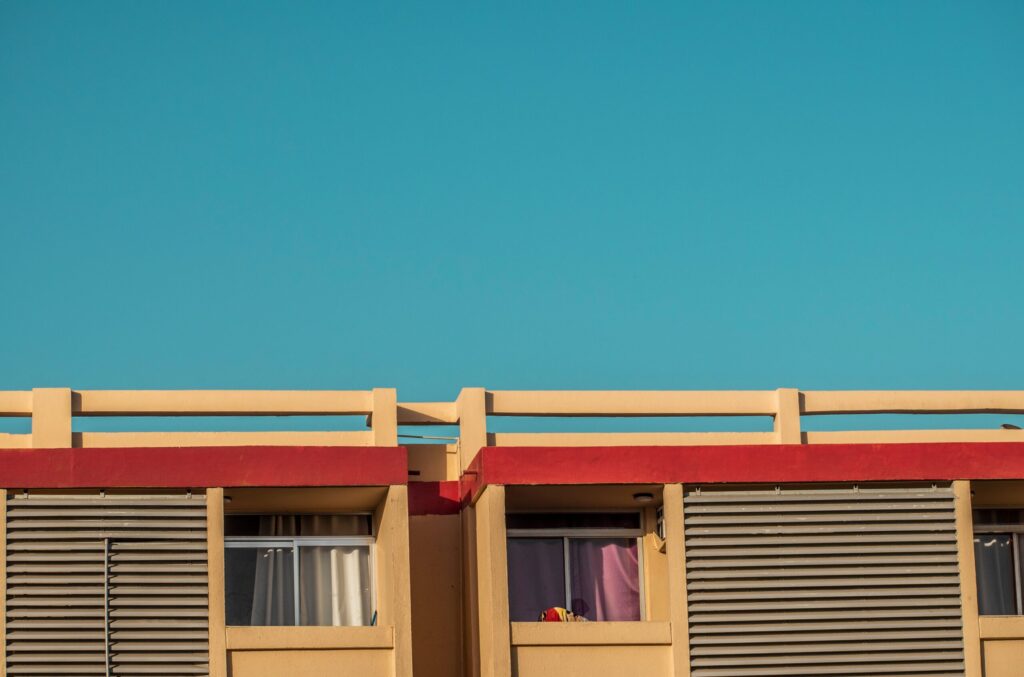 Minimalist crop of a cream-coloured Dubai mid-rise apartment facade with red trim and louvered shutters against a deep blue sky, representative of K One DubaiLand in DubaiLand Residence Complex