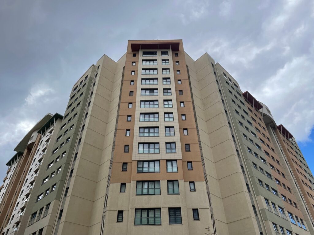 Low-angle view of a modern beige and orange residential tower against a cloudy sky, representative of National Bonds Oasis in Dubai Silicon Oasis, Dubai