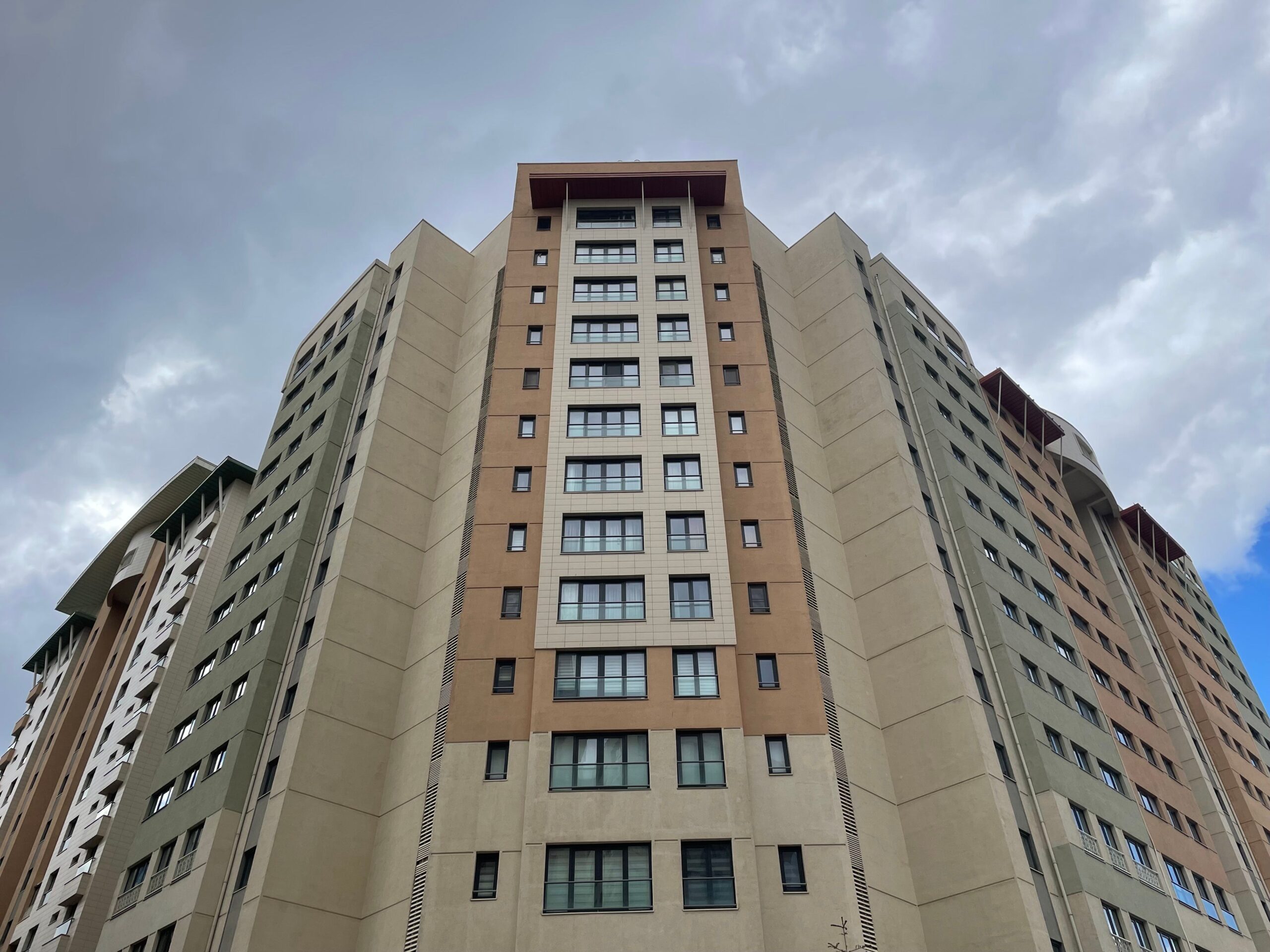 Low-angle view of a modern beige and orange residential tower against a cloudy sky, representative of National Bonds Oasis in Dubai Silicon Oasis, Dubai