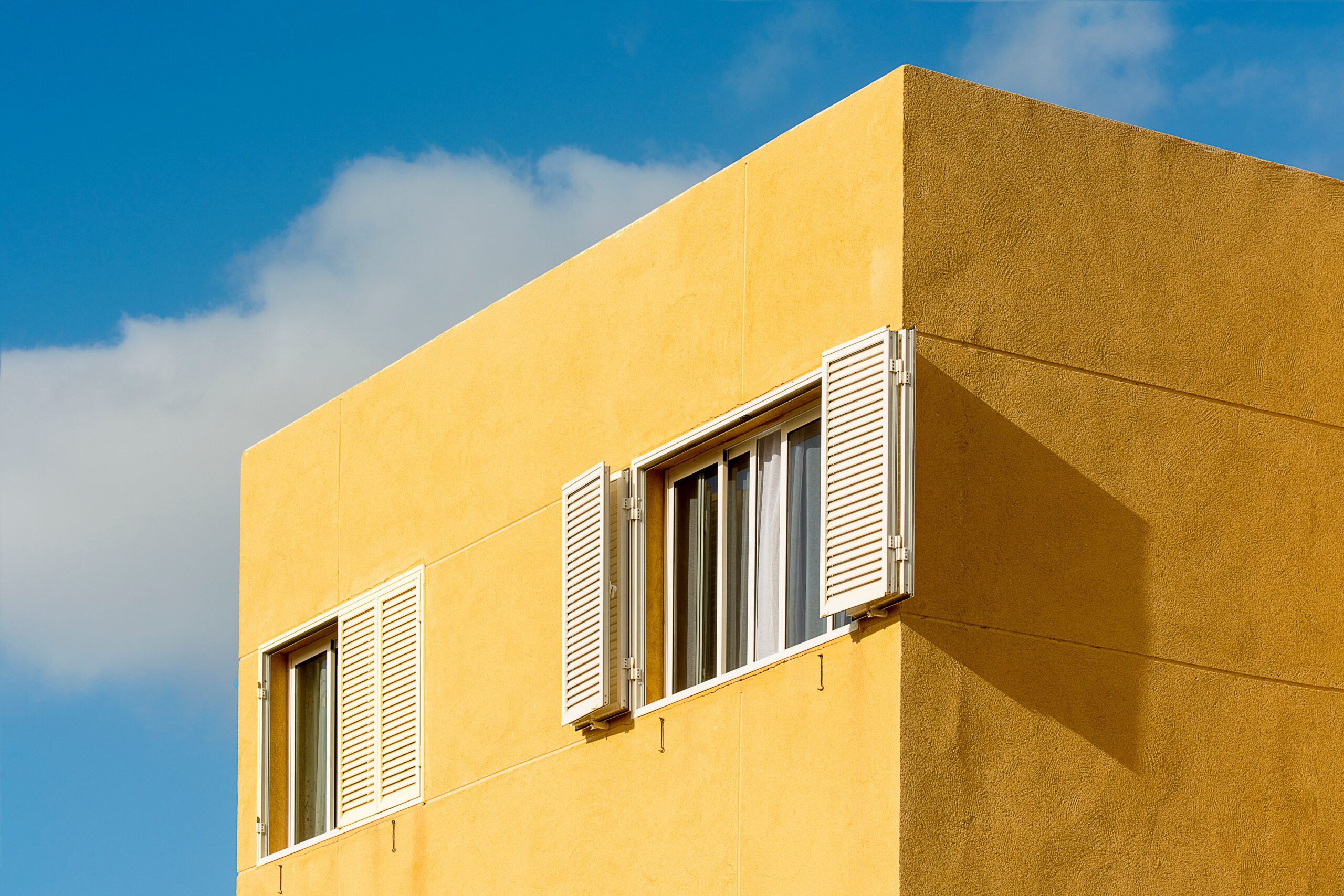 Corner of a modern mustard-yellow low-rise apartment building with white louvred window shutters against a clear blue sky in Dubai