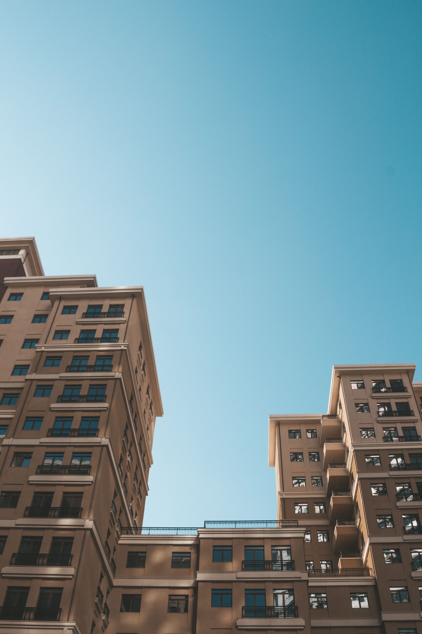 Low-angle view of a clean beige mid-rise Dubai apartment complex with balconies against a clear blue sky, representative of the Dubai Production City area near Qasr Sabah