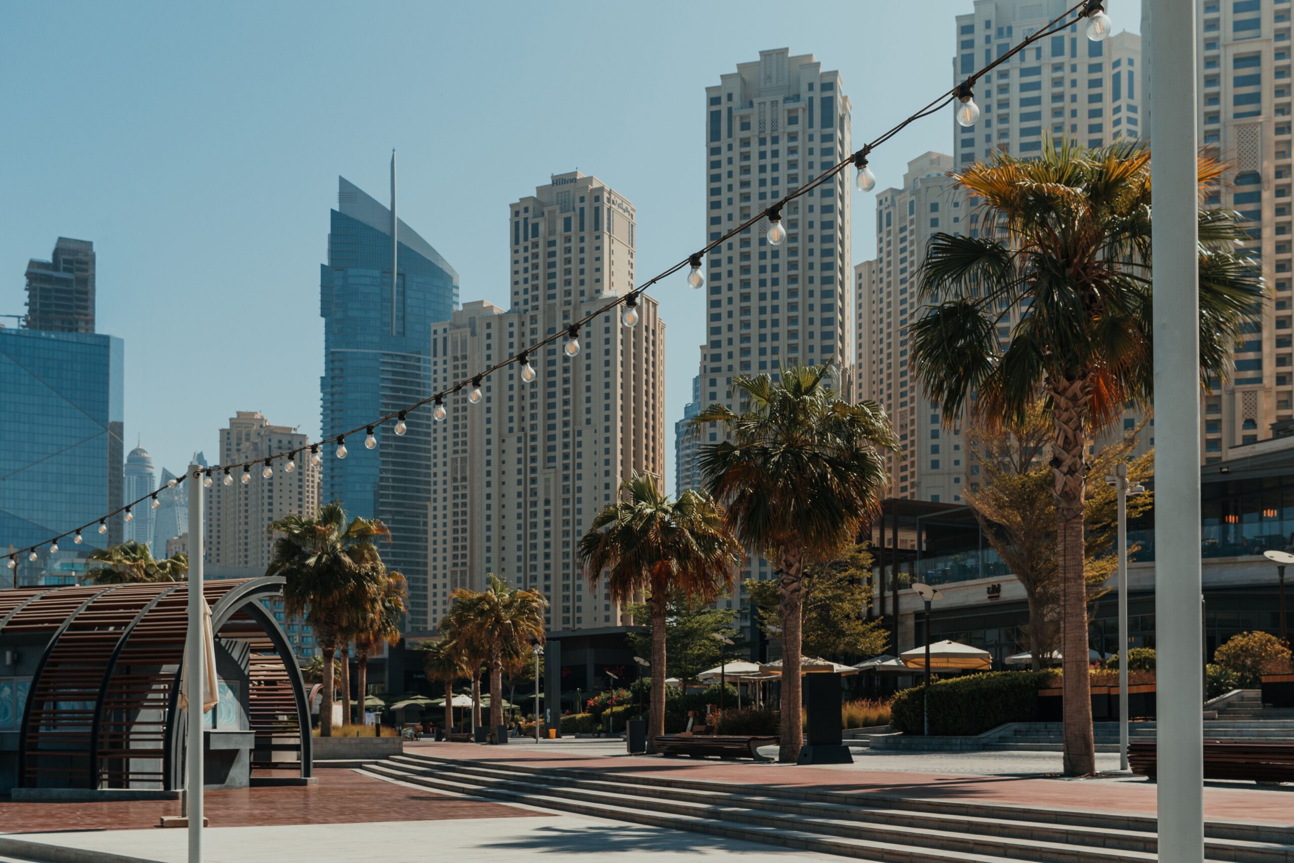 Dubai plaza and promenade lined with palm trees, string lights and a residential skyline in daylight, no people or legible signage visible