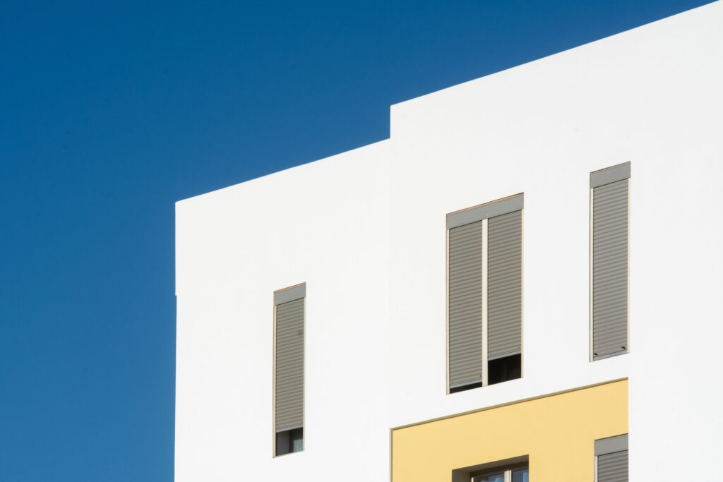 White residential building with grey shuttered windows and a yellow panel against a deep blue sky