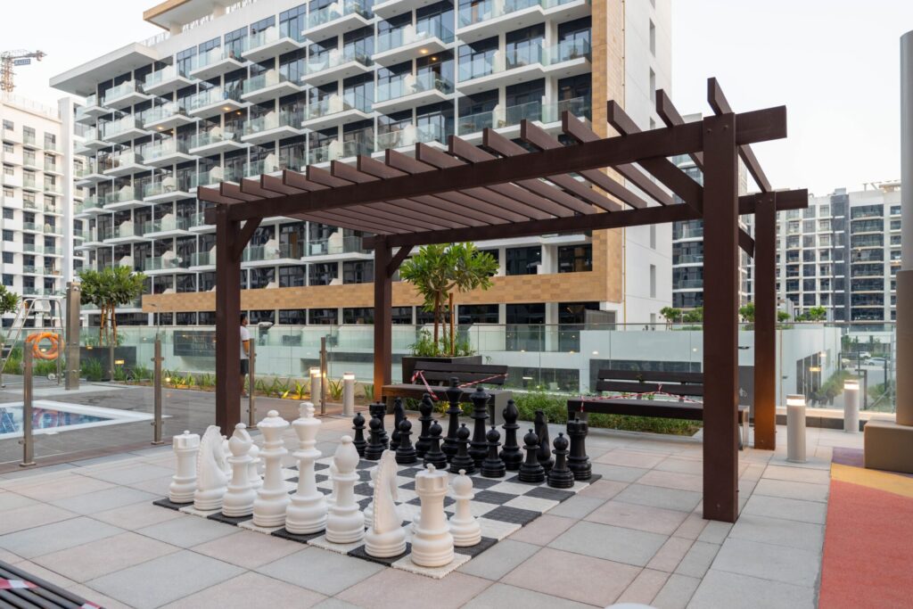 Rooftop community terrace with a wooden pergola and outdoor chess set, mid-rise residential buildings and a pool edge visible in the background